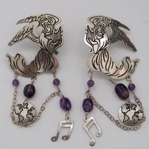 May include: A pair of silver-toned earrings featuring angel figures playing harps. Each earring has a chain with purple beads, a globe charm, and a musical note charm. The earrings are designed with intricate details.