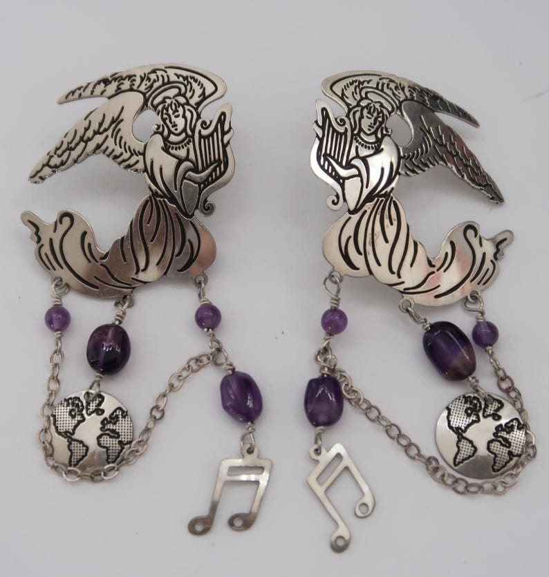 May include: A pair of silver-toned earrings featuring angel figures playing harps. Each earring has a chain with purple beads, a globe charm, and a musical note charm. The earrings are designed with intricate details.