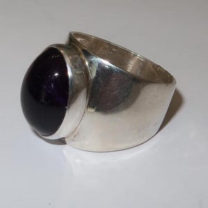 May include: A wide band ring crafted from polished silver metal, featuring a large, smooth, oval-shaped deep purple cabochon gemstone set securely in a bezel setting. The band has a thick, solid construction with a reflective, metallic finish.