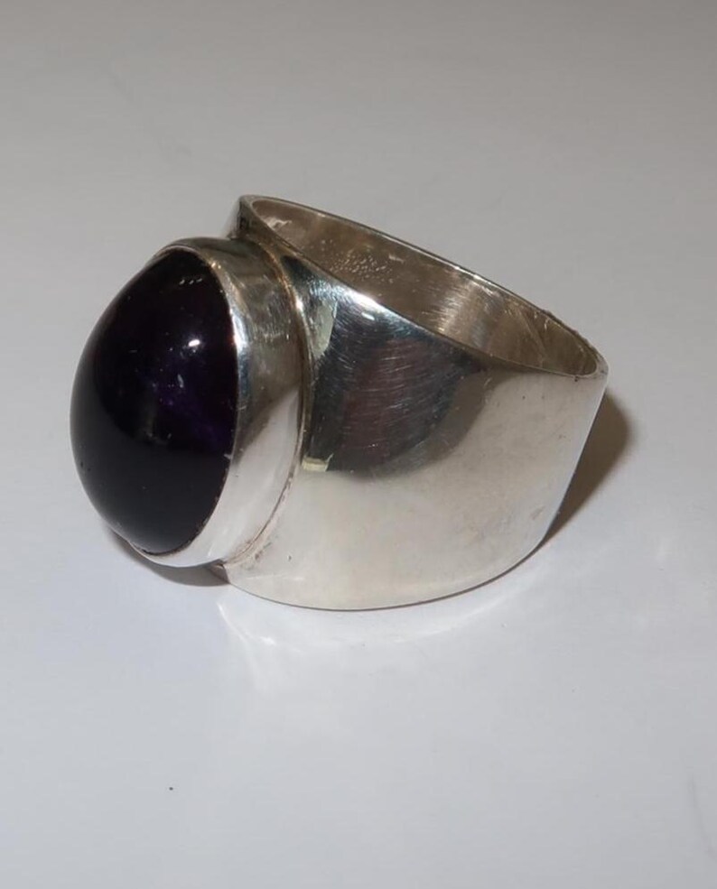 May include: A wide band ring crafted from polished silver metal, featuring a large, smooth, oval-shaped deep purple cabochon gemstone set securely in a bezel setting. The band has a thick, solid construction with a reflective, metallic finish.