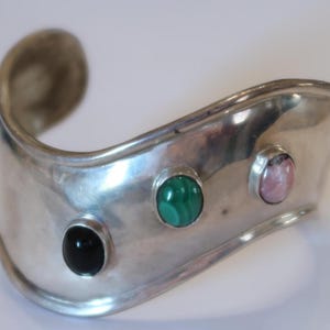 May include: A wide, curved silver cuff bracelet featuring three bezel-set cabochon gemstones arranged horizontally. The stones include a smooth black onyx, a vibrant green malachite with natural banding, and a soft pink rhodonite stone.