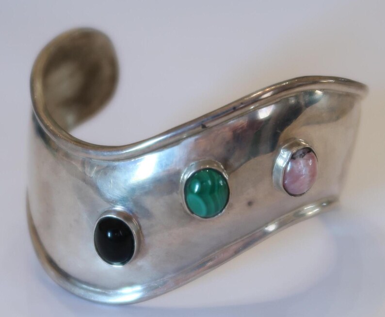 May include: A wide, curved silver cuff bracelet featuring three bezel-set cabochon gemstones arranged horizontally. The stones include a smooth black onyx, a vibrant green malachite with natural banding, and a soft pink rhodonite stone.