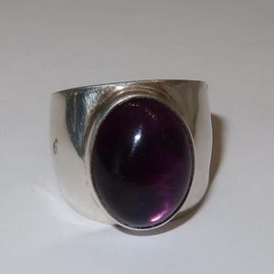 May include: A wide silver band ring featuring a large, smooth, oval-shaped deep purple cabochon gemstone set in the center. The metal surface of the ring has a polished, reflective finish with subtle brushed texture marks visible on the sides.