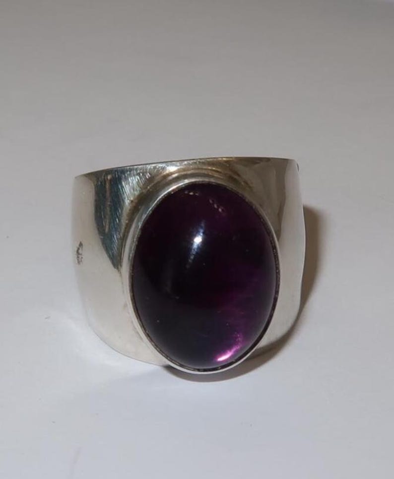 May include: A wide silver band ring featuring a large, smooth, oval-shaped deep purple cabochon gemstone set in the center. The metal surface of the ring has a polished, reflective finish with subtle brushed texture marks visible on the sides.