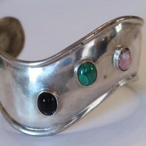 May include: A wide, polished silver cuff bracelet featuring a curved, wavy silhouette. The metal surface is set with three distinct oval cabochon gemstones: a solid black stone, a vibrant green banded stone, and a soft pink stone in a row.