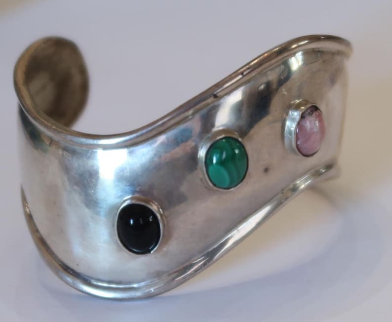 May include: A wide, polished silver cuff bracelet featuring a curved, wavy silhouette. The metal surface is set with three distinct oval cabochon gemstones: a solid black stone, a vibrant green banded stone, and a soft pink stone in a row.