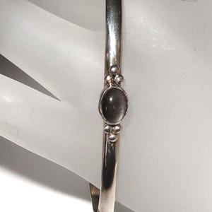 May include: A polished silver cuff bracelet featuring a smooth, oval-shaped dark gray stone set in the center. The stone is flanked by two small silver spheres on both the top and bottom, creating a symmetrical design on the slender metal band.
