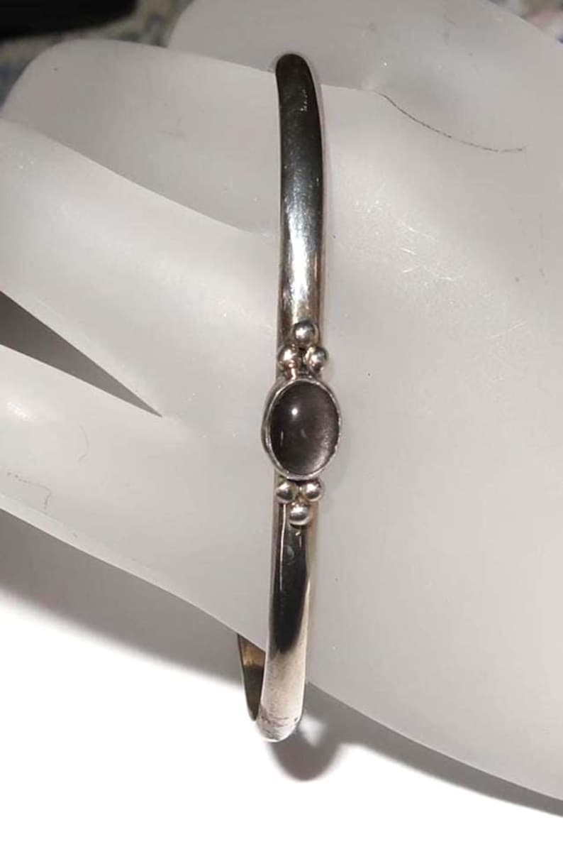 May include: A polished silver cuff bracelet featuring a smooth, oval-shaped dark gray stone set in the center. The stone is flanked by two small silver spheres on both the top and bottom, creating a symmetrical design on the slender metal band.