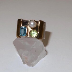 May include: A gold and silver ring featuring a rectangular blue gemstone, a round pearl, and a round green gemstone. The ring is set on a white surface.