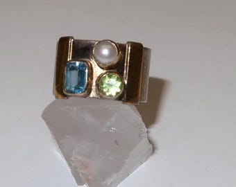 May include: A gold and silver ring featuring a rectangular blue gemstone, a round pearl, and a round green gemstone. The ring is set on a white surface.