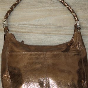 Puede incluir: Un bolso de mano de cuero marr&oacute;n con una correa trenzada y detalles plateados. El bolso tiene un brillo met&aacute;lico y una superficie ligeramente texturizada.