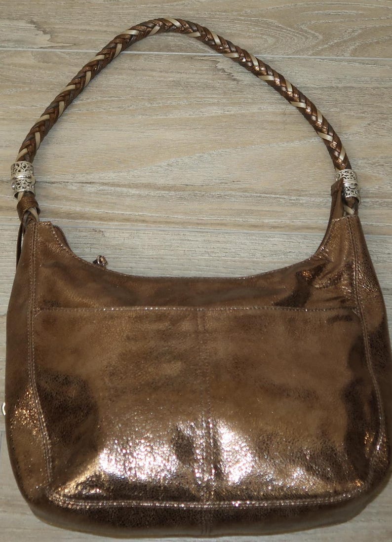 Puede incluir: Un bolso de mano de cuero marr&oacute;n con una correa trenzada y detalles plateados. El bolso tiene un brillo met&aacute;lico y una superficie ligeramente texturizada.