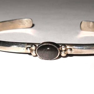May include: A silver-toned cuff bracelet featuring a smooth, polished metal band with an open back. The center of the bracelet is adorned with a single, oval-shaped dark gray stone set in a bezel, flanked by two small decorative metal spheres on each side.