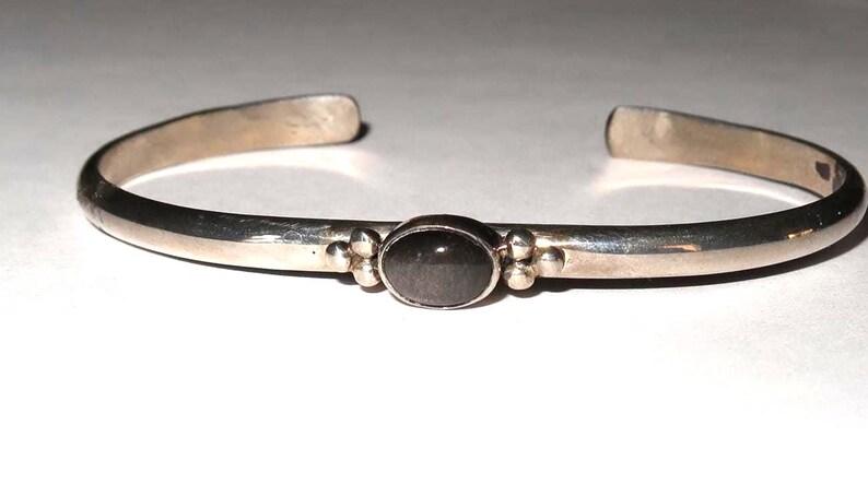 May include: A silver-toned cuff bracelet featuring a smooth, polished metal band with an open back. The center of the bracelet is adorned with a single, oval-shaped dark gray stone set in a bezel, flanked by two small decorative metal spheres on each side.