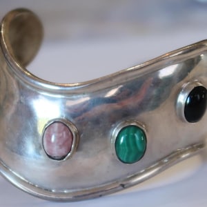 May include: A wide, polished silver cuff bracelet featuring a curved, undulating shape. The metal surface is set with three distinct oval cabochon gemstones: a light pink stone, a vibrant green malachite stone, and a smooth black stone.