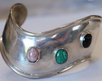 May include: A wide, polished silver cuff bracelet featuring a curved, undulating shape. The metal surface is set with three distinct oval cabochon gemstones: a light pink stone, a vibrant green malachite stone, and a smooth black stone.