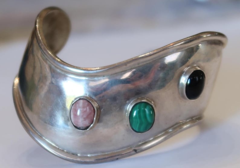 May include: A wide, polished silver cuff bracelet featuring a curved, undulating shape. The metal surface is set with three distinct oval cabochon gemstones: a light pink stone, a vibrant green malachite stone, and a smooth black stone.