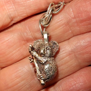 May include: Silver charm of a koala bear clinging to a branch. The charm is attached to a silver chain.