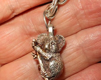 May include: Silver charm of a koala bear clinging to a branch. The charm is attached to a silver chain.