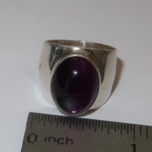 May include: A silver ring featuring a large, oval-shaped, deep purple cabochon gemstone set in the center. The ring is positioned next to a metal ruler that shows the band width is approximately 0.5 inches wide at the base of the setting.