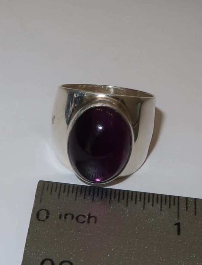 May include: A silver ring featuring a large, oval-shaped, deep purple cabochon gemstone set in the center. The ring is positioned next to a metal ruler that shows the band width is approximately 0.5 inches wide at the base of the setting.