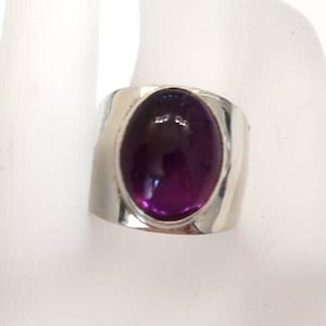 May include: A wide silver band ring featuring a large, smooth, oval-shaped deep purple gemstone set in the center. The metal band has a polished, reflective finish that curves around the finger, highlighting the contrast between the dark stone.
