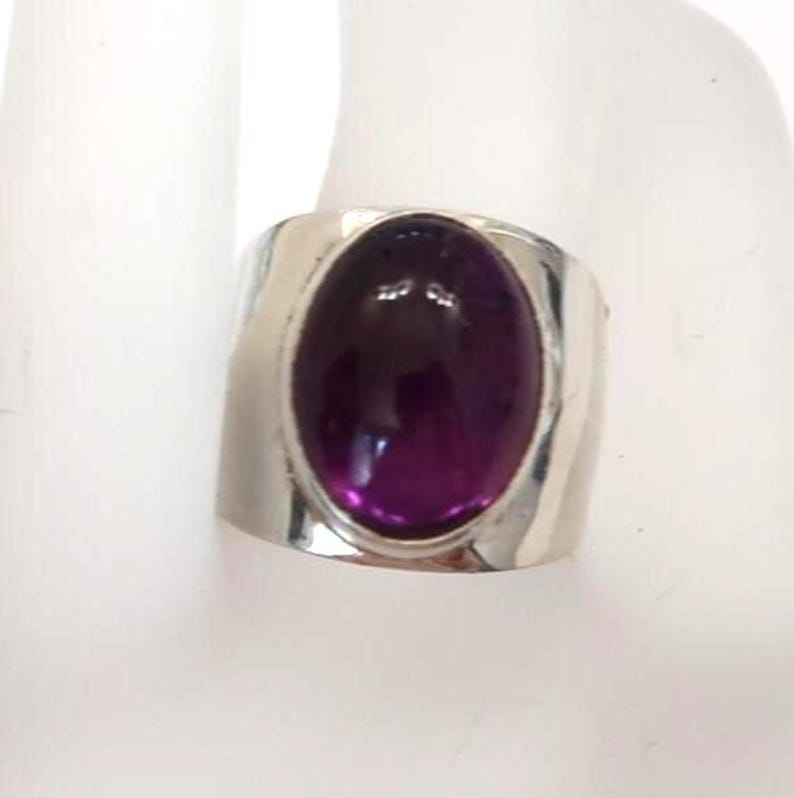 May include: A wide silver band ring featuring a large, smooth, oval-shaped deep purple gemstone set in the center. The metal band has a polished, reflective finish that curves around the finger, highlighting the contrast between the dark stone.
