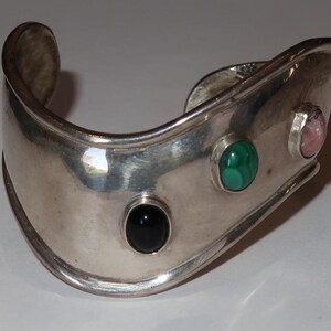 May include: A wide, polished silver cuff bracelet featuring three bezel-set cabochon gemstones arranged in a row. The stones include a smooth black onyx, a vibrant green malachite with natural banding, and a soft pink quartz or similar stone.