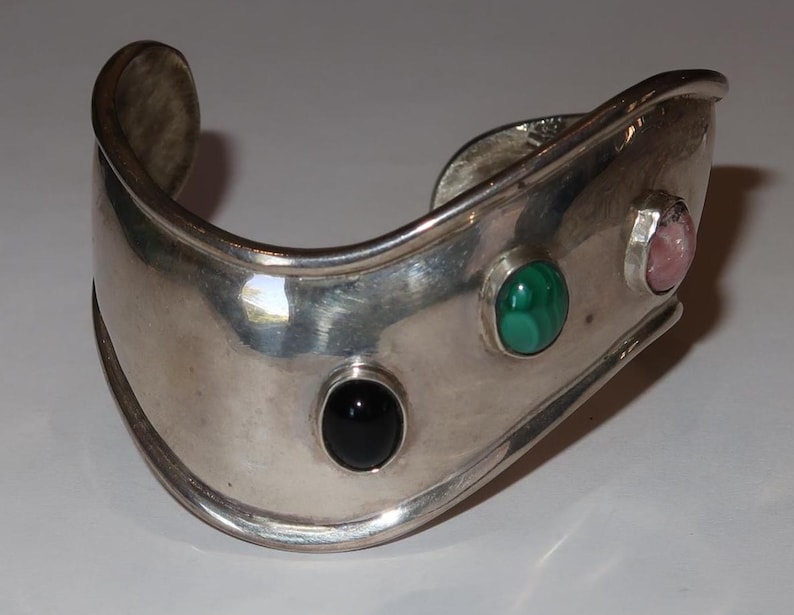 May include: A wide, polished silver cuff bracelet featuring three bezel-set cabochon gemstones arranged in a row. The stones include a smooth black onyx, a vibrant green malachite with natural banding, and a soft pink quartz or similar stone.