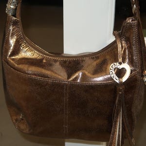 Puede incluir: Un bolso de mano de cuero marr&oacute;n con un dije de coraz&oacute;n plateado y una borla. El bolso tiene una cremallera y un asa superior.