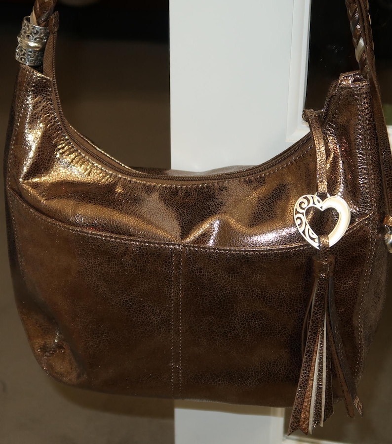 Puede incluir: Un bolso de mano de cuero marr&oacute;n con un dije de coraz&oacute;n plateado y una borla. El bolso tiene una cremallera y un asa superior.