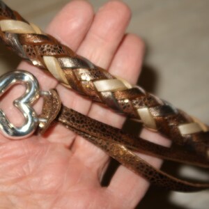 Puede incluir: Una correa de cuero marr&oacute;n con un dise&ntilde;o trenzado que presenta detalles marrones, blancos y dorados. Un dije de coraz&oacute;n plateado est&aacute; unido a la correa.