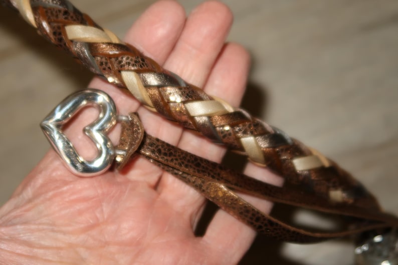 Puede incluir: Una correa de cuero marr&oacute;n con un dise&ntilde;o trenzado que presenta detalles marrones, blancos y dorados. Un dije de coraz&oacute;n plateado est&aacute; unido a la correa.