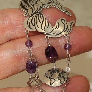 May include: A silver-toned pendant featuring an angel playing a harp, with dangling amethyst beads and a globe charm. The pendant has a musical note charm and is connected by a chain.