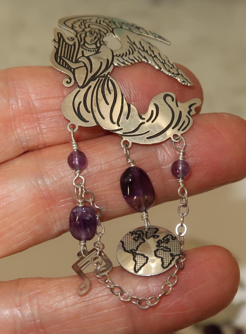 May include: A silver-toned pendant featuring an angel playing a harp, with dangling amethyst beads and a globe charm. The pendant has a musical note charm and is connected by a chain.