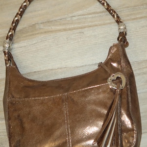 Puede incluir: Un bolso de mano de cuero marr&oacute;n con una correa trenzada y un dije de coraz&oacute;n plateado. El bolso tiene un brillo met&aacute;lico y una borla.