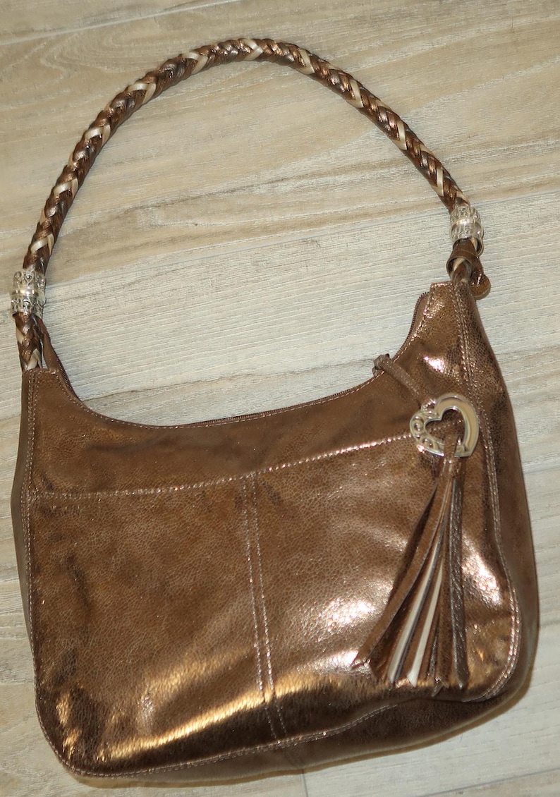Puede incluir: Un bolso de mano de cuero marr&oacute;n con una correa trenzada y un dije de coraz&oacute;n plateado. El bolso tiene un brillo met&aacute;lico y una borla.