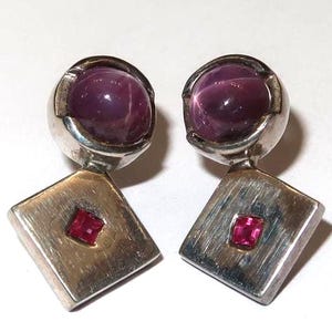 May include: A pair of silver earrings featuring a round, deep purple cabochon stone set in a circular frame above a diamond-shaped silver plate. Each plate has a brushed metal texture and is inlaid with a small, square-cut, vibrant red gemstone.