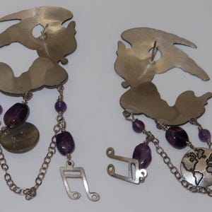 May include: A pair of silver-toned earrings with a whimsical design. Each earring features a stylized figure with dangling chains, purple beads, and decorative elements, including a musical note and a globe.