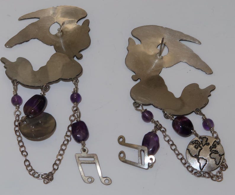 May include: A pair of silver-toned earrings with a whimsical design. Each earring features a stylized figure with dangling chains, purple beads, and decorative elements, including a musical note and a globe.