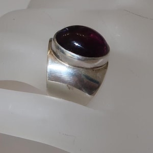 May include: A wide silver band ring featuring a large, smooth, oval-shaped deep purple cabochon gemstone set in a polished silver bezel. The metal surface of the ring is reflective and smooth, showcasing a solid construction with a rounded profile.