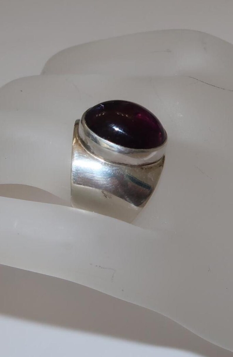 May include: A wide silver band ring featuring a large, smooth, oval-shaped deep purple cabochon gemstone set in a polished silver bezel. The metal surface of the ring is reflective and smooth, showcasing a solid construction with a rounded profile.