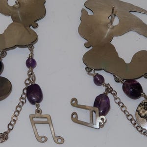 May include: Silver-toned earrings with a cloud-like top, dangling chains, and purple beads. One earring features a musical note charm, the other a globe. The earrings are approximately 3 inches long.