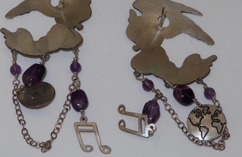 May include: Silver-toned earrings with a cloud-like top, dangling chains, and purple beads. One earring features a musical note charm, the other a globe. The earrings are approximately 3 inches long.