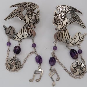 May include: A pair of silver-toned angel earrings. Each earring features an angel playing a harp, with dangling amethyst beads, a globe charm, and a musical note charm. The earrings have a detailed, ornate design.