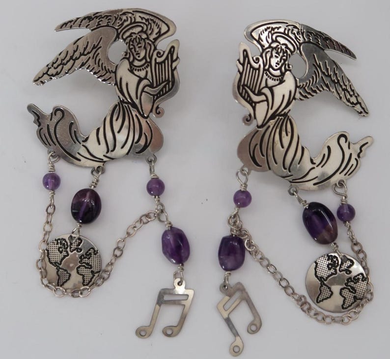 May include: A pair of silver-toned angel earrings. Each earring features an angel playing a harp, with dangling amethyst beads, a globe charm, and a musical note charm. The earrings have a detailed, ornate design.