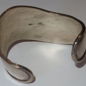 May include: A silver-toned cuff bracelet with a wide, open-ended band design. The metal surface features a brushed, matte finish with visible texture and slight variations in tone. The ends of the bracelet are rounded and smooth in appearance.