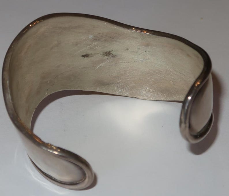 May include: A silver-toned cuff bracelet with a wide, open-ended band design. The metal surface features a brushed, matte finish with visible texture and slight variations in tone. The ends of the bracelet are rounded and smooth in appearance.