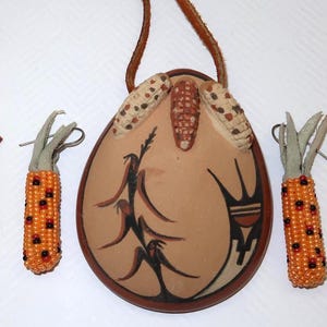 May include: A Native American-style jewelry set featuring a pendant and earrings. The pendant is a tan oval with painted designs and corn embellishments. The earrings are beaded corn shapes with silver hooks. The set includes a brown leather cord.