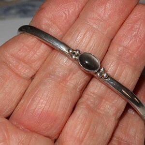 May include: A silver-toned metal bangle bracelet featuring a smooth, polished band. The center of the bracelet is adorned with a single oval-shaped dark gray stone, flanked on both sides by three small, decorative silver-toned metal spheres.